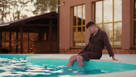 carefree-young-man-is-resting-in-spa-complex-with-outdoor-pool-surfing-internet-by-cellphone
